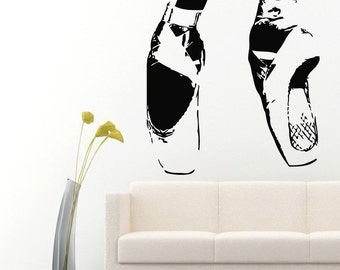 May include: Black and white silhouette wall decal of two ballet shoes. The shoes are positioned on a white wall with a white sofa in the background.