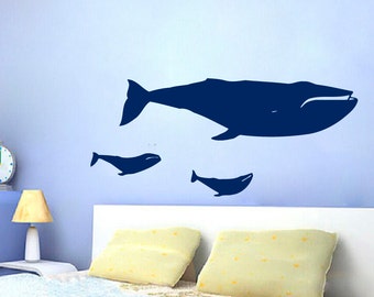 May include: A blue whale and two smaller whales are silhouetted on a light blue wall above a white headboard with two light yellow pillows.