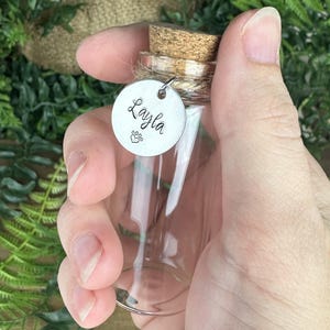 May include: A small, clear glass bottle with a cork stopper and a silver tag. The tag is engraved with the name "Layla" and a paw print. The bottle is held in a hand against a background of green foliage.