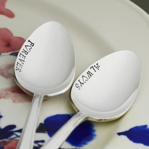 May include: Two silver spoons with the words "I love you" and "Forever" engraved on them. The spoons are on a white plate with a floral design.