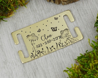 May include: A gold-colored metal tag with a floral and butterfly design. The tag has the name "Cleo" and a phone number. The tag has two curved cutouts on each end.