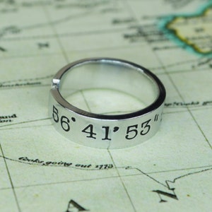 May include: A silver ring with the coordinates "56° 41' 53" engraved on it. The ring is lying on a map.