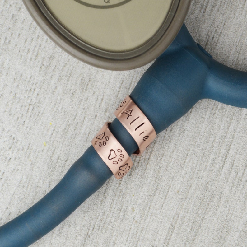 K&ouml;nnte beinhalten: Ein blaues Stethoskop mit zwei Kupferringen. Ein Ring tr&auml;gt den Namen "Allie" eingraviert und der andere Ring tr&auml;gt Pfotenabdr&uuml;cke eingraviert.