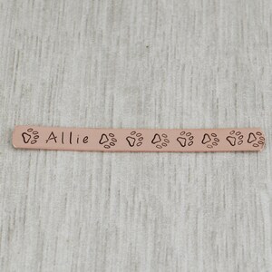K&ouml;nnte beinhalten: Ein personalisiertes Hundehalsband aus Kupfer mit dem eingravierten Namen "Allie". Das Halsband ist an den R&auml;ndern mit Pfotenabdr&uuml;cken verziert.
