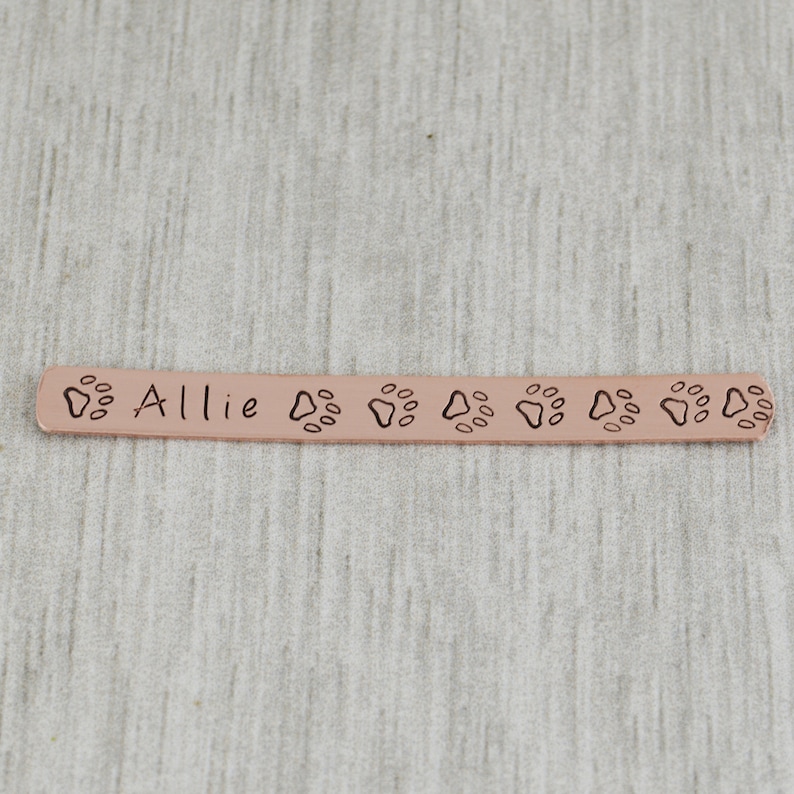 K&ouml;nnte beinhalten: Ein personalisiertes Hundehalsband aus Kupfer mit dem eingravierten Namen "Allie". Das Halsband ist an den R&auml;ndern mit Pfotenabdr&uuml;cken verziert.