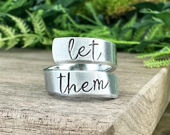 May include: A silver ring with the words "let them" engraved on it. The ring is made of two bands that wrap around each other.