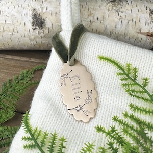 Puede incluir: Un calcetín de Navidad personalizado con una etiqueta festoneada que dice "Ellie". La etiqueta es de metal de color claro y está adornada con ilustraciones botánicas. El calcetín es blanco y tiene una cinta de terciopelo verde.