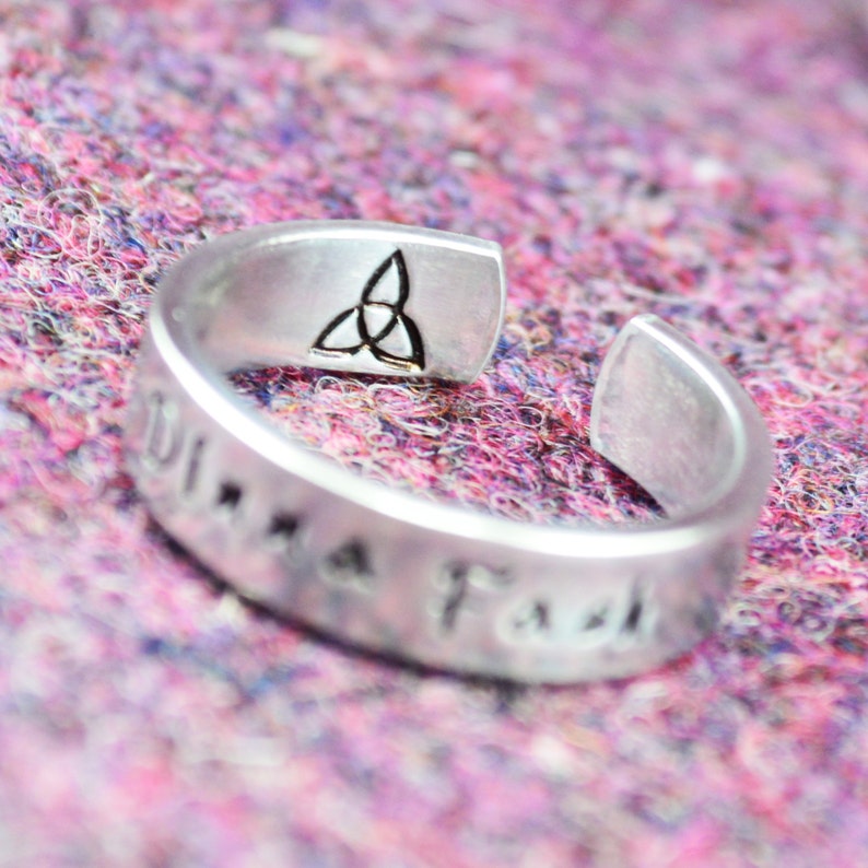 May include: Silver adjustable ring featuring a Celtic knot design and the inscription "Una F&aacute;s" on the band.