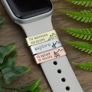 Könnte beinhalten: Drei goldene, silberne und roségoldene Uhrenarmbänder mit gravierten Bergszenen und Text. Das obere Band trägt die Aufschrift "The Mountains Are Calling". Das mittlere Band trägt die Aufschrift "explore". Das untere Band trägt die Aufschrift "I'd Rather Be Hiking".