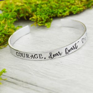 Könnte beinhalten: Silbernes Armreif-Armband mit der Inschrift "COURAGE, liebes Herz" und zwei kleinen Herzen.