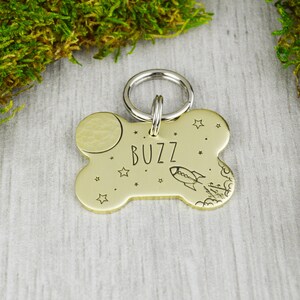 Könnte beinhalten: Ein goldfarbener, knochenförmiger Haustieranhänger mit dem eingravierten Namen "BUZZ" in der Mitte. Der Anhänger zeigt ein Mond- und Raketendesign mit Sternen. Ein silberner Ring ist angebracht.