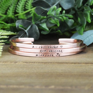May include: Three stacked rose gold bracelets with the inscription "a-choidhche gun aithoras misneach" in Gaelic script.