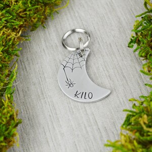 May include: A silver-coloured, crescent-shaped pet tag featuring a spider web and spider design. The word "KILO" is printed on the tag. A silver ring is attached for easy attachment to a pet's collar.