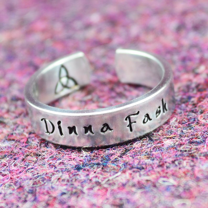 May include: A silver ring with a Celtic knot design and the inscription "Dinna Fask" on a pink and purple fuzzy surface.