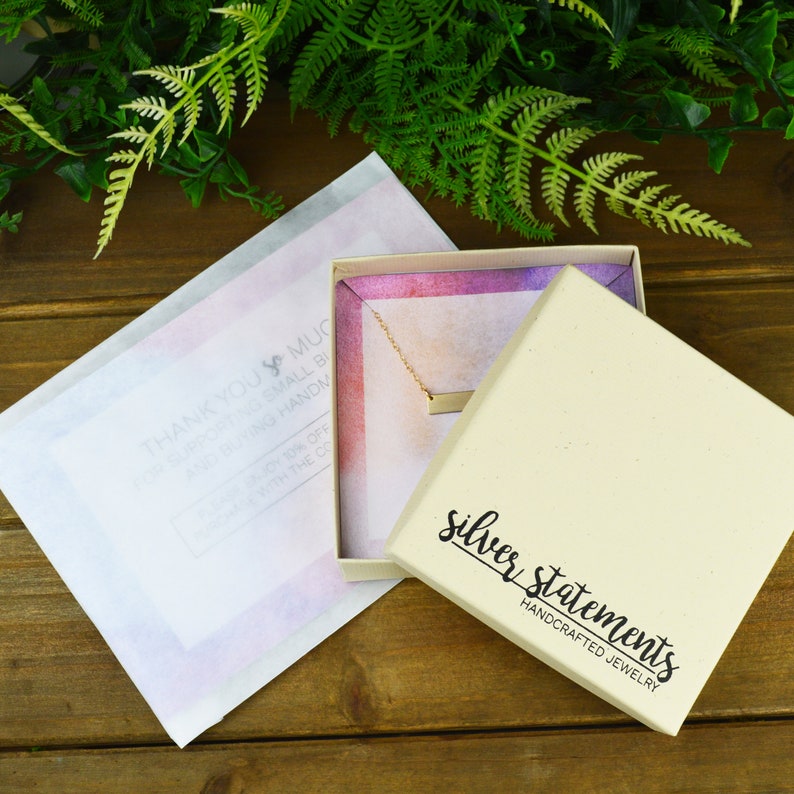 K&ouml;nnte beinhalten: Eine kleine, wei&szlig;e Schachtel mit einer goldenen Bar-Halskette darin. Die Schachtel hat innen ein rosafarbenes und violettes Aquarelldesign und auf der Unterseite steht "silver statements handcrafted jewelry" gedruckt.