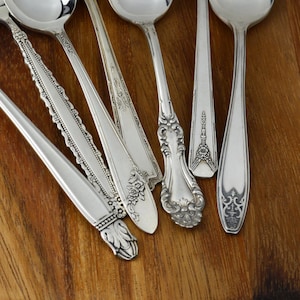 May include: Six silver spoons with ornate designs on the handles. The spoons are arranged on a wooden surface.