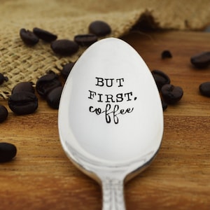 May include: A silver spoon with the phrase "But first, coffee" engraved on the front. The spoon is surrounded by coffee beans.