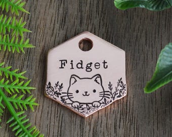Op de afbeelding: Een koperkleurige, zeshoekige dierenpenning met de naam "Fidget" in vette, zwarte letters. Een schattige kattenillustratie staat onder de naam, omringd door bloemmotieven. De penning heeft een gat aan de bovenkant om te bevestigen.