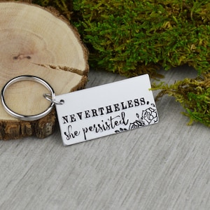 Könnte beinhalten: Ein silberner rechteckiger Schlüsselanhänger mit den Worten "NEVERTHELESS, she persisted" in schwarzer Schrift. Auf der rechten Seite befindet sich ein Blumendesign. Der Schlüsselanhänger ist an einem silbernen Ring befestigt. Der Hintergrund umfasst eine Holzscheibe und grünes Moos.