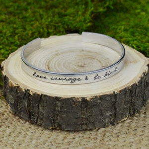 May include: A silver cuff bracelet with the engraved words "have courage & be kind." The bracelet is displayed on a small, round wooden slice with a natural bark edge. The background features green moss.