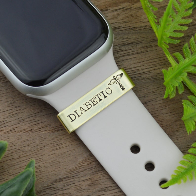 K&ouml;nnte beinhalten: Eine wei&szlig;e Apple Watch mit einem goldenen Metallband, auf dem "DIABETIC" mit einem medizinischen Symbol steht. Das Band ist mit einem wei&szlig;en Band an der Uhr befestigt.