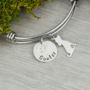 May include: A silver bangle bracelet with a charm that says "Gunther" and features a paw print inside a heart. A small silver dog silhouette charm hangs from the bangle.