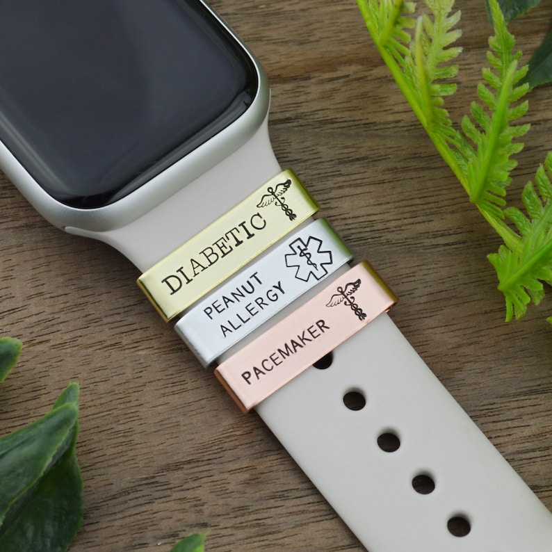 K&ouml;nnte beinhalten: Drei goldene, silberne und ros&eacute;goldene medizinische Alarm-Armb&auml;nder f&uuml;r eine Smartwatch. Die B&auml;nder haben die Worte "Diabetic", "Peanut Allergy" und "Pacemaker" eingraviert. Jedes Band hat auch ein medizinisches Symbol eingraviert.