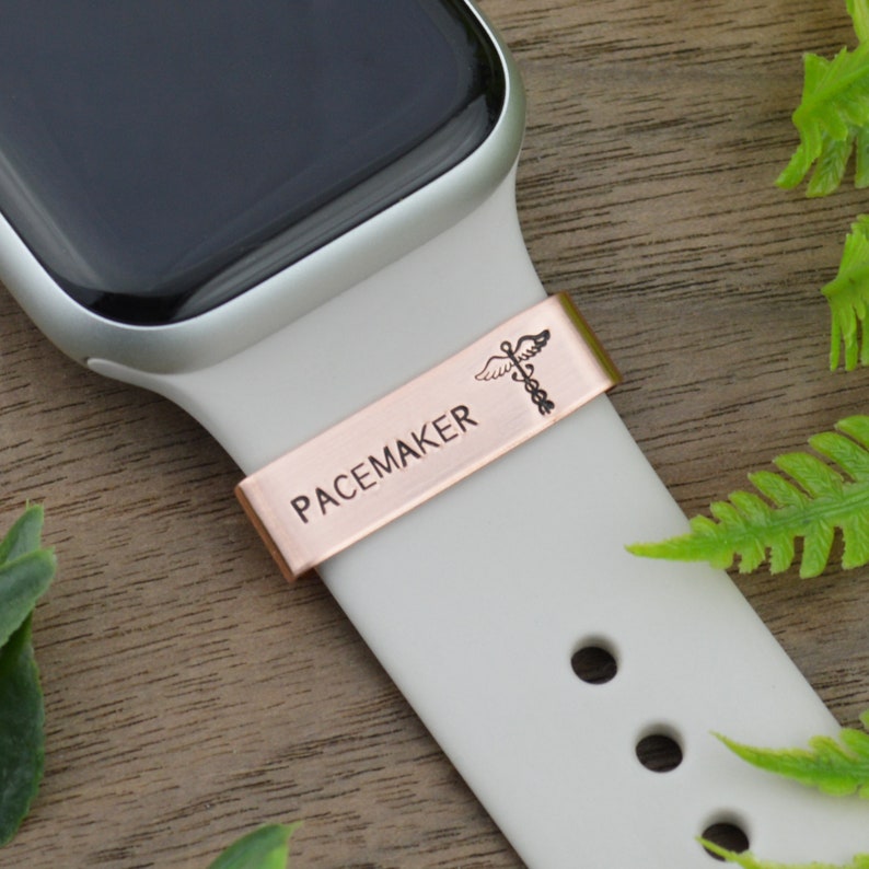 K&ouml;nnte beinhalten: Eine wei&szlig;e Apple Watch mit einem ros&eacute;goldenen Armband und einem medizinischen Warnhinweis mit der Aufschrift "PACEMAKER" und einem Caduceus-Symbol.