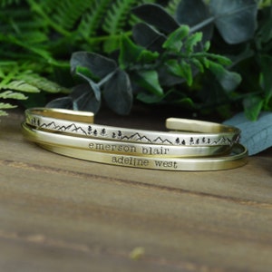 May include: Three gold bracelets stacked on top of each other. The top bracelet has a mountain range and trees design. The middle bracelet has the text "emerson blair" engraved on it. The bottom bracelet has the text "adeline west" engraved on it.