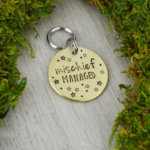 Peut inclure: Une médaille ronde dorée pour animaux de compagnie avec l'inscription "mischief managed" en script noir, entourée de petites étoiles. La médaille est attachée à un anneau fendu argenté. Le fond comprend de la mousse verte.