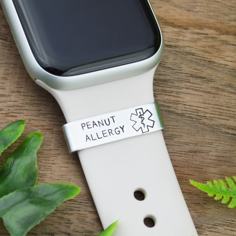 K&ouml;nnte beinhalten: Eine wei&szlig;e Apple Watch mit einem silbernen Armband und einem medizinischen Warnband mit der Aufschrift "Erdnussallergie" und einem medizinischen Symbol.
