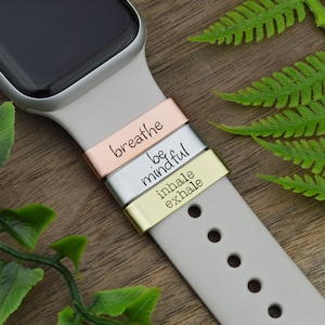 Puede incluir: Una banda de reloj Apple blanca con tres bandas metálicas adjuntas. Las bandas están grabadas con las palabras "breathe", "be mindful" y "inhale exhale".