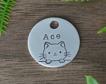 Op de afbeelding: Een ronde, zilveren dierenpenning met een gat aan de bovenkant. De penning toont de naam "Ace" boven een cartoon kat illustratie. De kat heeft een lachend gezicht en kijkt over een lijn.