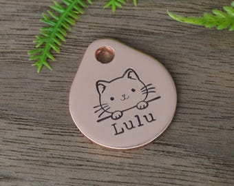 Op de afbeelding: Een roségouden huisdierpenning met een kattenillustratie en de naam "Lulu" gegraveerd. De penning heeft een druppelvorm met een gat voor bevestiging. De achtergrond is een houten oppervlak met groene bladeren.