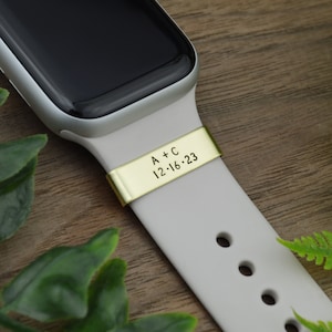 Könnte beinhalten: Weißes Apple Watch Armband mit einem goldenen Metallband, das mit "A + C 12.16.23" graviert ist.
