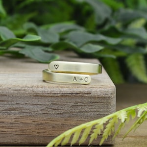 May include: A gold-toned, open-ended ring with a heart symbol engraved on one side and the initials "A+C" on the other side. The ring is on a wooden surface.