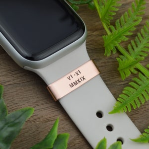 Könnte beinhalten: Ein roségoldener Uhrenarmbandverschluss mit den eingravierten römischen Ziffern VI-XI MMXIX. Der Verschluss ist an einem hellgrauen Uhrenarmband befestigt. Das Zifferblatt der Uhr ist schwarz. Farne sind im Hintergrund.