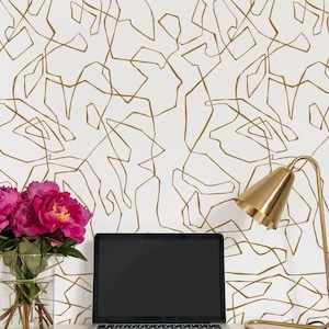 May include: A white desk with a gold lamp, a laptop computer, and a vase of pink flowers. The wall behind the desk is covered in white wallpaper with a gold geometric pattern.