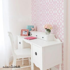 May include: A white desk with four drawers and a white chair. The desk is against a pink wall with a white geometric pattern. There is a small elephant figurine on the desk.