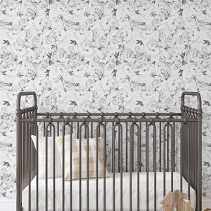 May include: A white crib with a black metal frame and a white mattress. The crib is decorated with a black and white floral wallpaper. A rocking horse and a suitcase are in the foreground.
