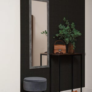 May include: A full-length rectangular mirror with a gray frame hangs on a dark textured wall. A small brown leather handbag, sunglasses, and a vase with greenery sit on a black table. A gray velvet stool and a pair of orange heels complete the decor.