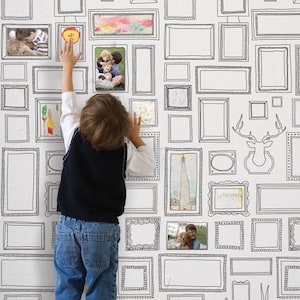 May include: A child in a dark vest and blue jeans reaches towards a wall covered in framed artwork and photos. The wall features a repeating pattern of black outlined frames, some filled with colorful drawings and family photos.