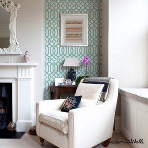 May include: A white armchair with a floral patterned pillow sits in a living room with a teal and white geometric patterned wallpaper. A framed picture with a colorful design hangs on the wall.