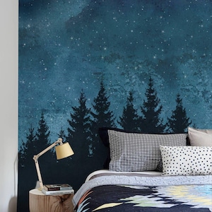 Könnte beinhalten: Ein Wandbild mit einem dunkelblauen Nachthimmel voller weißer Sterne über einer Silhouette von Kiefern. Die Szene befindet sich hinter einem Bett mit einer gemusterten Steppdecke, einem karierten Kissen und einem hölzernen Nachttisch mit Lampe.