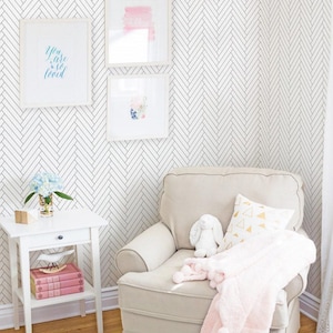 Puede incluir: Un sillón blanco con una manta rosa y una almohada dorada se encuentra en una habitación con paredes blancas y un papel tapiz de espiga en blanco y negro. Una mesita auxiliar blanca con un cajón y libros se encuentra junto al sillón. La pared tiene dos marcos blancos con obras de arte.