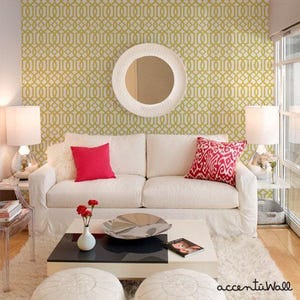 May include: A white couch with red and white pillows sits in front of a wall with a gold geometric pattern wallpaper. A round mirror hangs above the couch. A coffee table with a black top and white base sits in front of the couch. There are two white poufs in front of the coffee table.