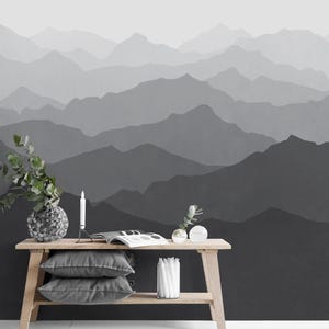 Op de afbeelding: Een houten tafel met twee planken, met een grijze vaas met groene bladeren, een witte kaars, een tijdschrift, twee heldere glazen bollen en twee grijze kussens. De tafel staat tegen een muur met een zwart-grijze bergketen behang.