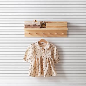 Op de afbeelding: Een wit houten plank met haken, een klein knuffeldier en gevouwen kleding. Een witte jurk met een bloemenpatroon hangt aan een hanger onder de plank. Twee witte schoenen staan op de vloer onder de jurk.