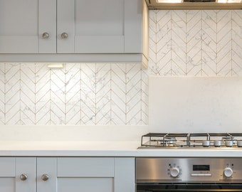 May include: A white kitchen with gray cabinets and a stainless steel oven and stovetop. The backsplash is white marble with gold chevron patterns.