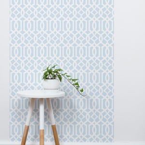 May include: A white side table with three wooden legs sits in front of a light blue wall with a white geometric pattern. A small potted plant with green leaves sits on the table.